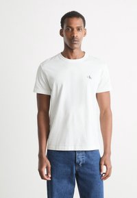 Hvid bomulds t-shirt med rund halsudskæring og korte ærmer. Har et diskret logo på brystet. Bærer blå denim jeans.
