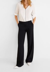 Blouse blanche avec passepoil noir, manches longues retroussées, associée à un pantalon noir à jambes larges et des chaussures blanches à bouts pointus. Design simple.