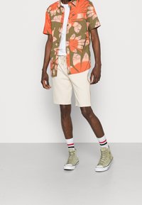 Kortærmet skjorte i orange og olivenfarvet med blomstertryk; hvid t-shirt under. Cremefarvede shorts og olivenfarvede high-top sneakers.