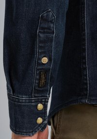 Rękaw ciemnoniebieskiej denimowej koszuli, ozdobiony dwoma okrągłymi złotymi guzikami i metką z tkaniny, z widocznym szwem i detalami wywiniętego mankietu.