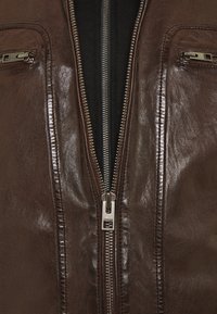 Giacca di pelle marrone con una zip centrale in argento, tasca con zip, texture liscia e dettagli cuciti lungo le cuciture.