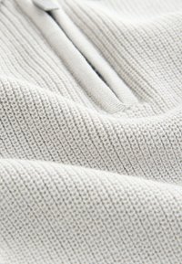 Hellgrauer, strukturierter Stoff mit einem gerippten Muster und einem weichen, gewebten Aussehen. Am oberen Rand befindet sich ein kurzes Reißverschlussdetail.