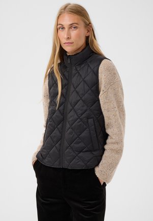 Femme aux longs cheveux blonds porte un gilet matelassé noir sur un pull en maille beige et un pantalon noir, debout les mains dans les poches devant un fond clair.