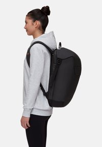 Sac à dos noir avec des bretelles rembourrées, un grand compartiment et une poche latérale, porté par une personne en sweat à capuche gris devant un fond uni.