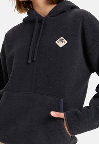 Sweat à capuche en polaire noire avec une poche avant, une capuche réglable et un patch logo Roxy. Texture douce et coupe décontractée.