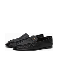 Et par svarte skinnloafers med flettet kant og et lite gulldekorativt emblem på overflaten, vist mot en hvit bakgrunn.