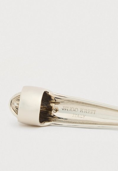 Λευκόχρυσος γυαλιστερός παπουτσόκουτσος με λείο, επιμηκυμένο σχέδιο· διαθέτει στρογγυλεμένο άκρο και μεταλλικό καπάκι κοντά στη λαβή. Σημειωμένος με τη φράση «Hugo Kreit Italy».