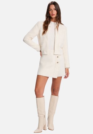 Femme aux longs cheveux bruns portant une veste crème texturée, une jupe assortie avec des boutons dorés, un haut blanc côtelé et des bottes crème jusqu'aux genoux.