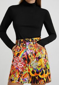 Donna che indossa un maglione a collo alto nero a maniche lunghe e una gonna colorata con stampa di draghi, fiori, fiamme e creature mitologiche, con le mani in tasca.