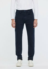 Jeans de mezclilla azul oscuro con un diseño de pierna recta, que presentan un estilo clásico de cinco bolsillos y detalles de costura sutiles. Se llevan con una camiseta blanca de manga larga.