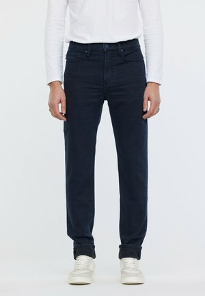 Jeans de mezclilla azul oscuro con un diseño de pierna recta, que presentan un estilo clásico de cinco bolsillos y detalles de costura sutiles. Se llevan con una camiseta blanca de manga larga.