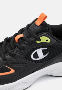 Scarpa sportiva nera con tomaia in mesh e sintetico, dettagli arancioni e verdi, logo prominente e suola bianca, caratterizzata da motivi testurizzati.