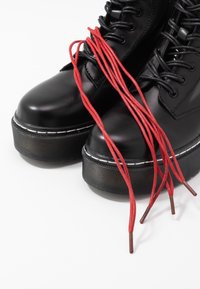 Bottes à plateforme noires avec semelles épaisses, lacets noirs et longs lacets rouges drapés sur le devant sur un fond blanc.