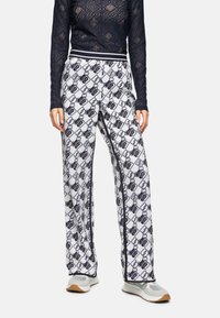 Weiße gemusterte Hose mit einem schwarzen Kettendesign. Elastischer Bund mit schwarz-weißen Streifen, kombiniert mit hellgrauen Sneakers.