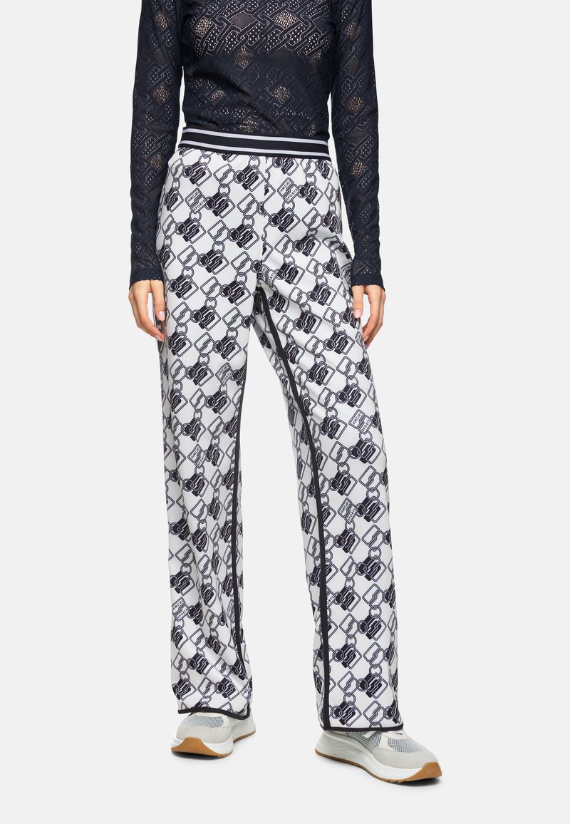 Weiße gemusterte Hose mit einem schwarzen Kettendesign. Elastischer Bund mit schwarz-weißen Streifen, kombiniert mit hellgrauen Sneakers.