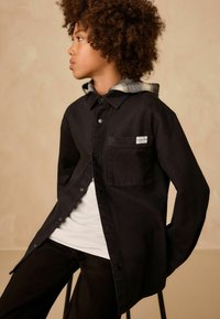 Schwarzes Hemd mit Knöpfen, einer Brusttasche und sichtbaren Nähten, über einem weißen T-Shirt getragen und mit einem grauen karierter Kapuzenpullover kombiniert.