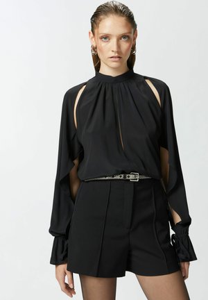 Blusa nera con maniche lunghe e fluide e dettagli a taglio, abbinata a pantaloni corti neri a vita alta e una cintura metallica. Tessuto morbido.