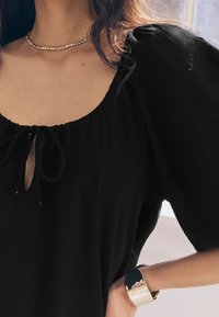 Femme portant un haut noir à manches bouffantes avec un petit nœud devant, un collier de perles et un large bracelet en argent à son poignet.