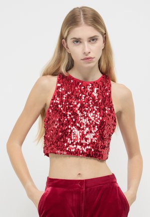 Woman with long blonde hair wearing a sleeveless red sequin crop top and red velvet pants with hands in pockets against a plain background.