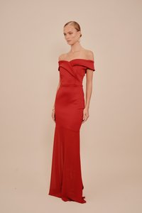 Nő, hátul fésült hajjal, váll nélküli, testhez simuló piros ruhában, átlátszó, hosszú szoknyával, bézs háttér előtt áll.