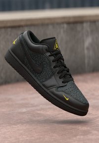 Svart low-top sneakers med teksturerte mørkegrå overlegg og gule detaljer. Inkluderer svarte lisser og en gummisåle for grep.