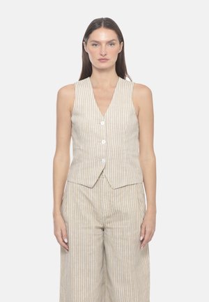 Donna che indossa un gilet smanicato a righe verticali beige e bianche e pantaloni larghi abbinati, in piedi su uno sfondo semplice.
