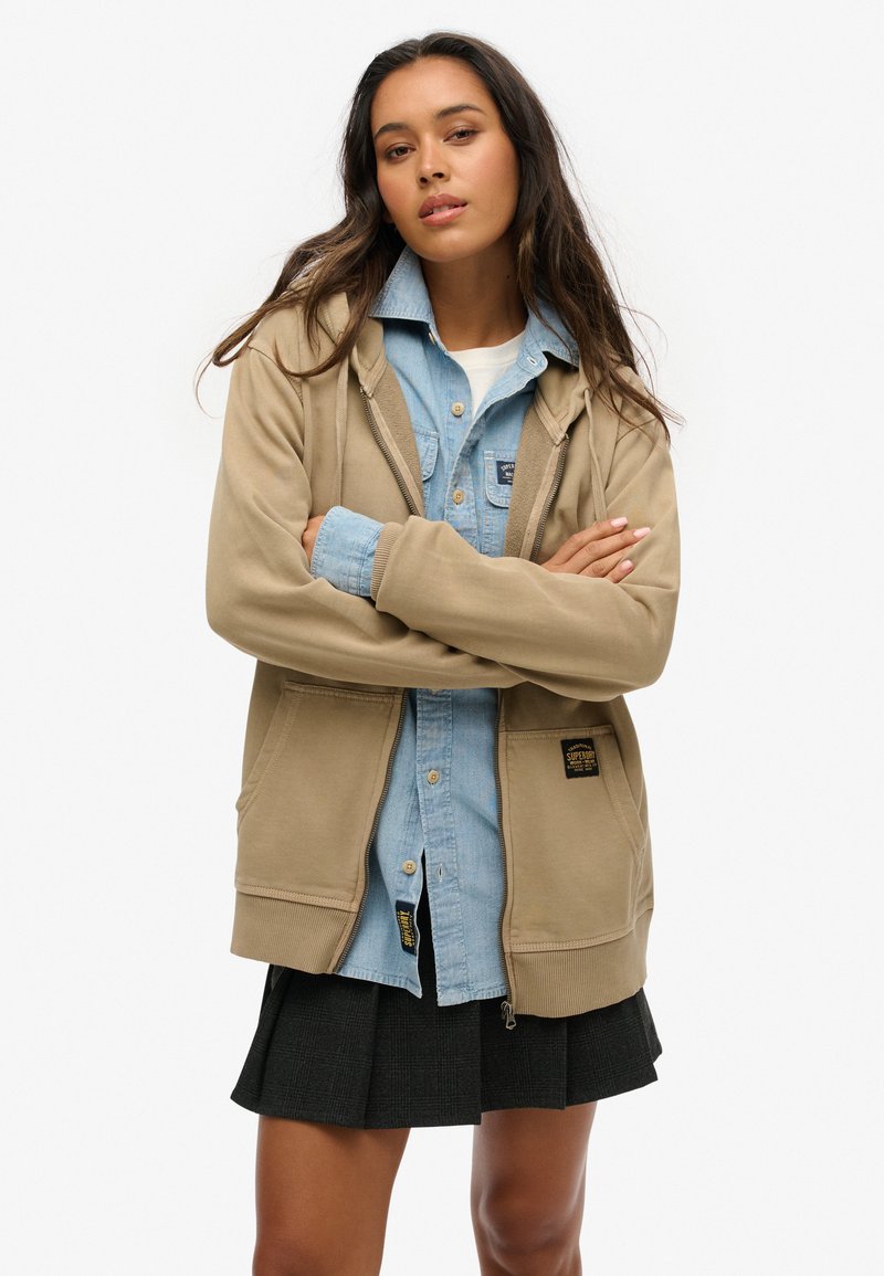 Hoodie beige zippé avec poches avant, superposé sur une chemise en denim bleu clair, associé à une jupe plissée noire. Texture de tissu lisse.