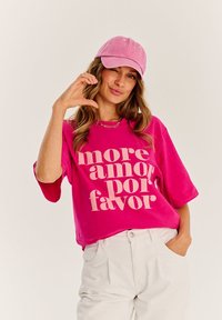 Růžové oversized tričko s nápisem "more amor por favor" v krémové barvě, doplněné o světle růžovou kšiltovku. Bílé kalhoty s bočními kapsami.