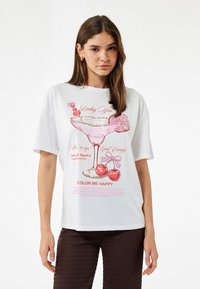 Witte T-shirt met korte mouwen, voorzien van een graphic van een roze cocktailglas met kersen en de tekst "Lucky You" en "COLOR ME HAPPY." Zachte stof, ontspannen pasvorm.