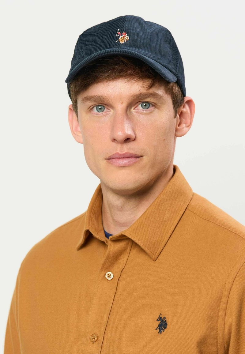 Jeune homme aux yeux bleus portant une casquette noire avec un logo brodé et une chemise marron boutonnée avec un petit logo sombre sur la poitrine.