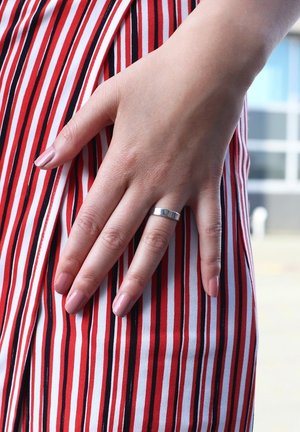 Zilveren bandring op een hand met lichtroze nagels, rustend op een rood-wit gestreepte stof. De ring heeft een gladde, gepolijste oppervlakte.