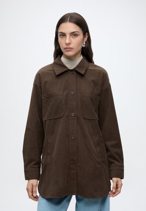 Kvinde iført en brun kordfløjlsjakke med store brystlommer over en lys rullekrave og blå jeans mod en hvid baggrund.