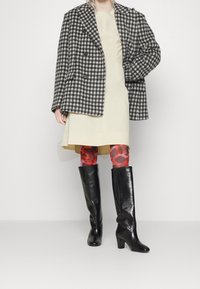 Blazer de cuadros en blanco y negro de corte oversize, vestido crema, mallas rojas con patrón, botas de cuero negras hasta la rodilla con tacón grueso.