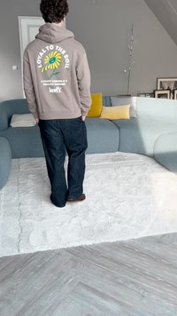 Sudadera marrón claro con texto gráfico y diseño de flores, combinada con jeans azul oscuro y zapatos marrones, en un interior moderno con un sofá curvado.