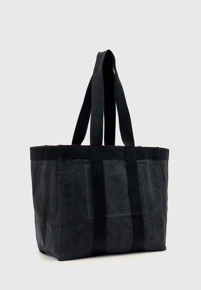 Sac fourre-tout en toile noire avec deux longues anses et coutures renforcées, debout sur un fond gris clair uni.