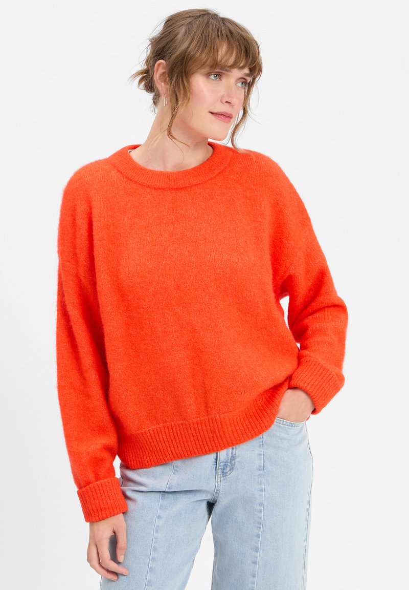 Svetlo oranžna prevelika blagovna svise, izdelana iz mehkega pletenega materiala, s krožno ovratnikom in rebrastimi manšetami, v kombinaciji s svetlo modrimi kavbojkami.