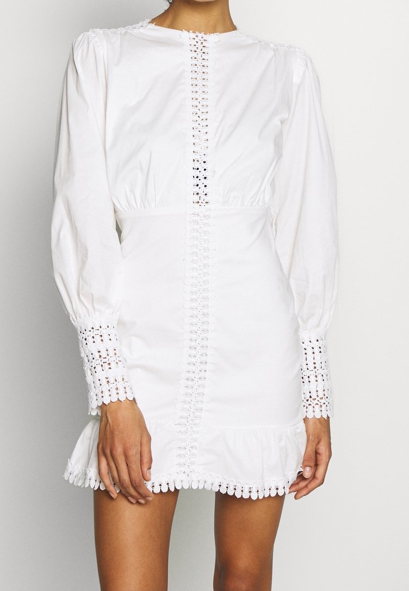 Robe courte blanche à manches longues avec bordure en dentelle au centre, aux poignets et à l'ourlet, avec une taille froncée et un léger volant en bas.
