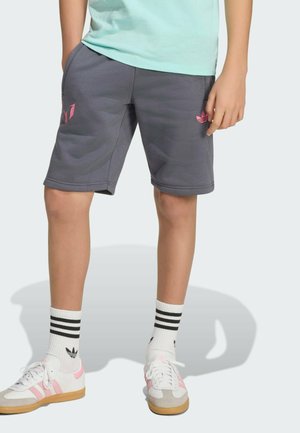 Dítě nosí šedé šortky Adidas s růžovými logy, bílé ponožky Adidas s černými pruhy a bílé tenisky s růžovými akcenty.