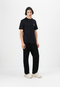 Sort bomulds t-shirt med korte ærmer, der har et minimalistisk grafik på brystet. Parret med sorte bukser og beige sneakers.