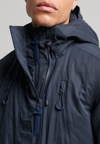 Granatowa wodoodporna kurtka z wysokim kołnierzem, regulowanymi sznurkami i wieloma kieszeniami zapinanymi na suwak. Posiada detal z logo na suwaku.