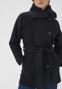 Námornícky modrý kabát s opaskom vyrobený z mäkkej látky, s vysokým golierom, tromi gombíkmi a dvoma prednými vreckami.