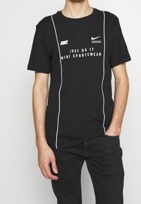 Schwarzes Baumwoll-T-Shirt mit kurzen Ärmeln, weißen Akzenten und Nike-Branding, einschließlich des "Just Do It"-Textes und Logos auf der Vorderseite.