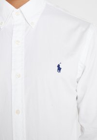 Biała koszula zapinana na guziki z klasycznym kołnierzykiem, długimi rękawami oraz granatowym haftowanym logo polo gracza na lewej piersi.