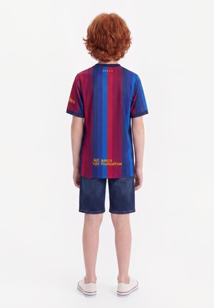 Kind mit roten Haaren steht mit dem Rücken zur Kamera, trägt ein blau-rot gestreiftes Barcelona Foundation Shirt, dunkle Shorts und weiße Turnschuhe vor einfarbigem Hintergrund.