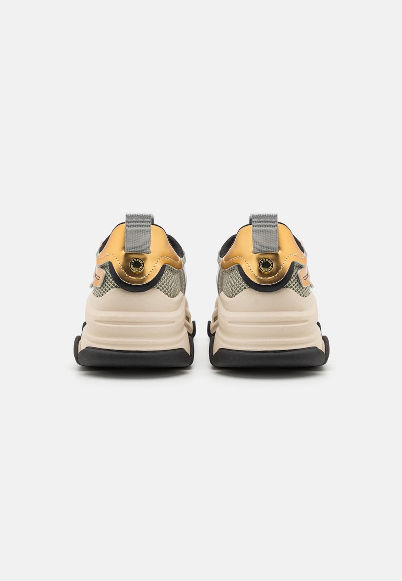 Zapatillas beige y amarillas con un cuerpo de malla texturizada, suela de goma negra y una prominente pestaña plateada en el talón. Presenta acentos de herrajes.