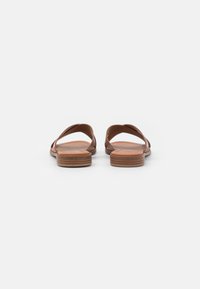 Des sandales en cuir marron présentent des brides entrecroisées et une semelle plate avec une texture superposée subtile. Aucune ornementation ni motif visible.