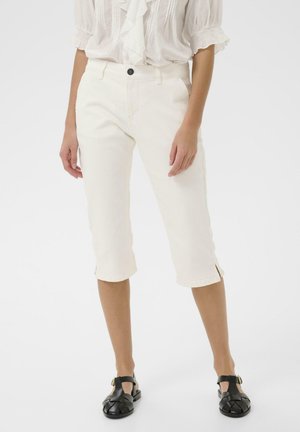 Žena má na sebe biele krátke nohavice s bočnými rozparkami, doplnené bielou blúzkou a čiernymi sandálmi s uzavretou špičkou a remienkami okolo členkov.