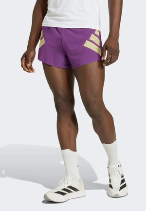 Athlet trägt violette Laufshorts mit beigen Streifen, weiße Crew-Socken und weiße Sneakers mit schwarzen Streifen vor einem schlichten Hintergrund.