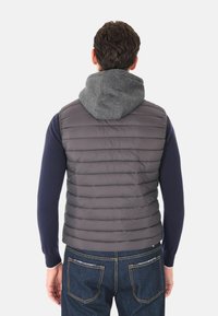 Uomo che indossa un gilet imbottito grigio con cappuccio grigio sopra una maglietta a maniche lunghe blu navy e jeans blu, mostrato di spalle.