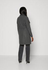 Vrouw met lang, donker haar in een grijze jas met riem, donkergrijze jeans en zwarte hoge hakken, staand met haar rug naar de camera op een effen achtergrond.